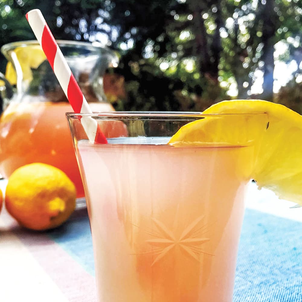 A glass of our low-sugar all-natural lavender lemonade garnished with a lemonade sitting in front of a pitcher and lemons