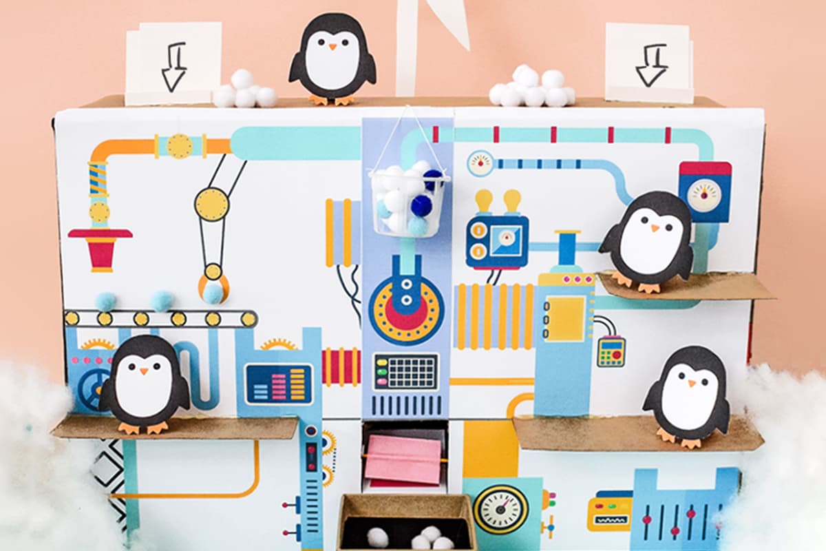 Our snowball factory kids craft sitting against a peach and light blue background surrounded by fake snow with cardboard penguins standing on different levels