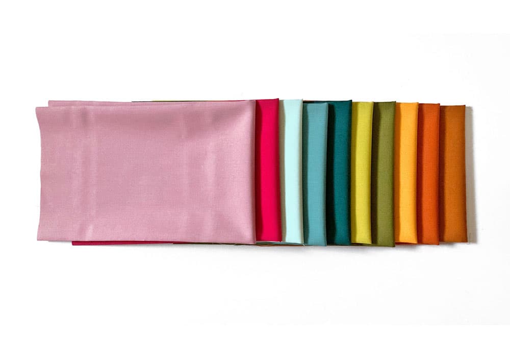 Ten fat quarters of colorful poplin used to make our soft village kids playset folded and sitting on a white background
