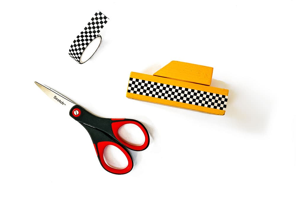 A wood taxi cab sitting on a white background next to checkered tape and scissors