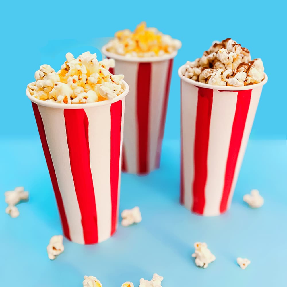 three cups of popcorn sprinkled with our homemade popcorn seasoning mixes sitting in front of a blue background