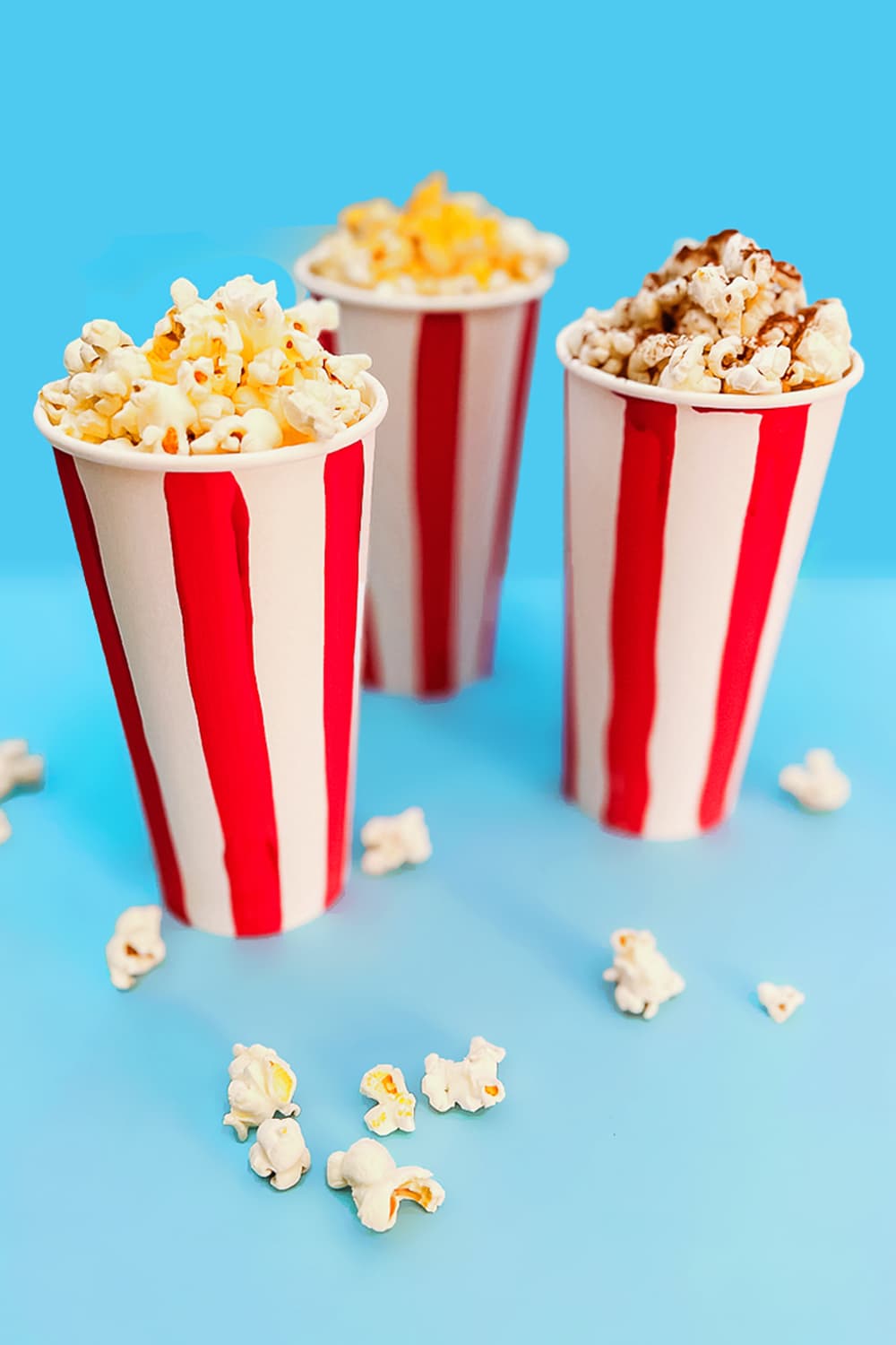 three cups of popcorn sprinkled with our diy popcorn seasonings sitting in front of a blue background