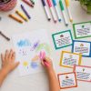 A young child's hands drawing a colorful crayon scene on white paper, surrounded by scattered crayons, bright markers, and a pink polka dot crayon holder. Five illustrated Summer Creative Prompt Cards in green, teal, blue, yellow, and coral are fanned out to the right, each showing a different open-ended activity challenge.