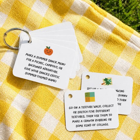 A flat lay of blank white square activity cards on a colorful patterned blanket on green grass. Several cards are threaded onto a silver book ring, fanned out to show the hole punch in the upper right corner, with a few loose cards scattered nearby. Bright natural daylight and vibrant colors.