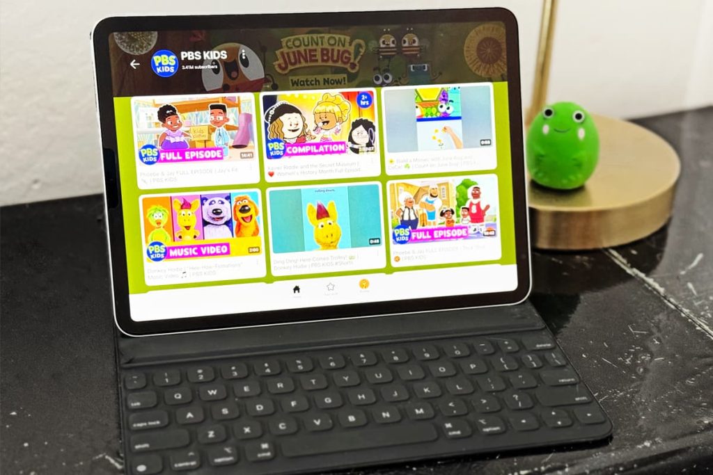 A tablet sitting on a desk next to a lamp and children's toys. The YouTube Kids app is on the screen of the tablet.