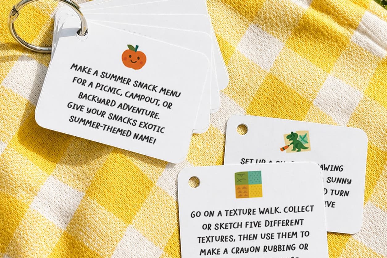 A flat lay of blank white square activity cards on a colorful patterned blanket on green grass. Several cards are threaded onto a silver book ring, fanned out to show the hole punch in the upper right corner, with a few loose cards scattered nearby. Bright natural daylight and vibrant colors.